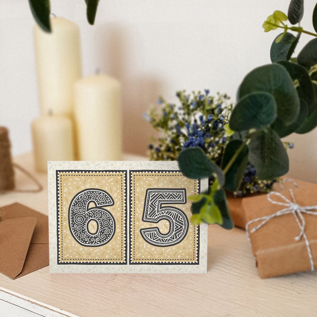 Gold 65th birthday card sat on a wooden surface with candles, plants and gifts.