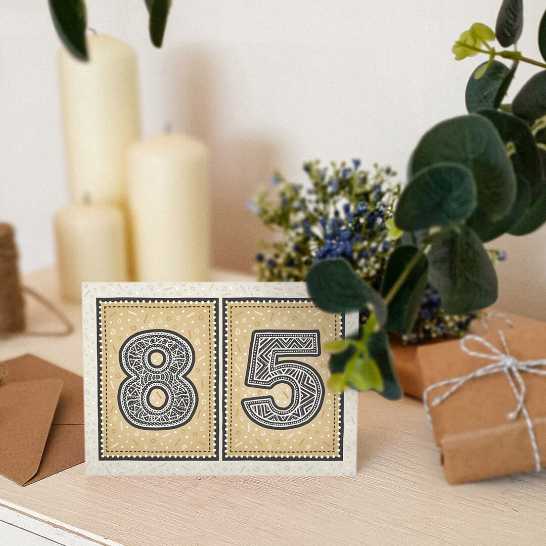 Gold 85th birthday card stood on a cream wooden table with candles, plants and gifts.