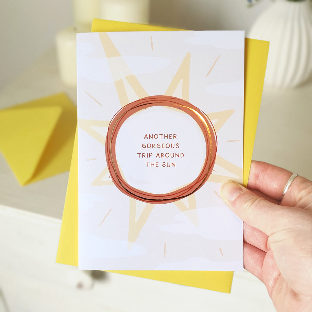 Birthday card with sun design and text held by a hand, with a yellow envelope.