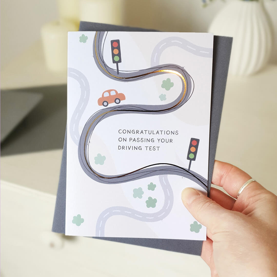 Hand holding a card with a road and car illustration and text 'Congratulations on passing your driving test'.