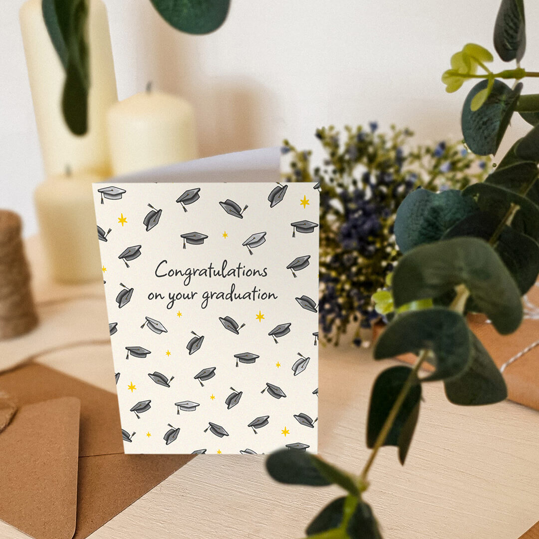 unique graduation card featuring illustrated mortarboards sat on a cream wooden dresser surrounded by plants and candles