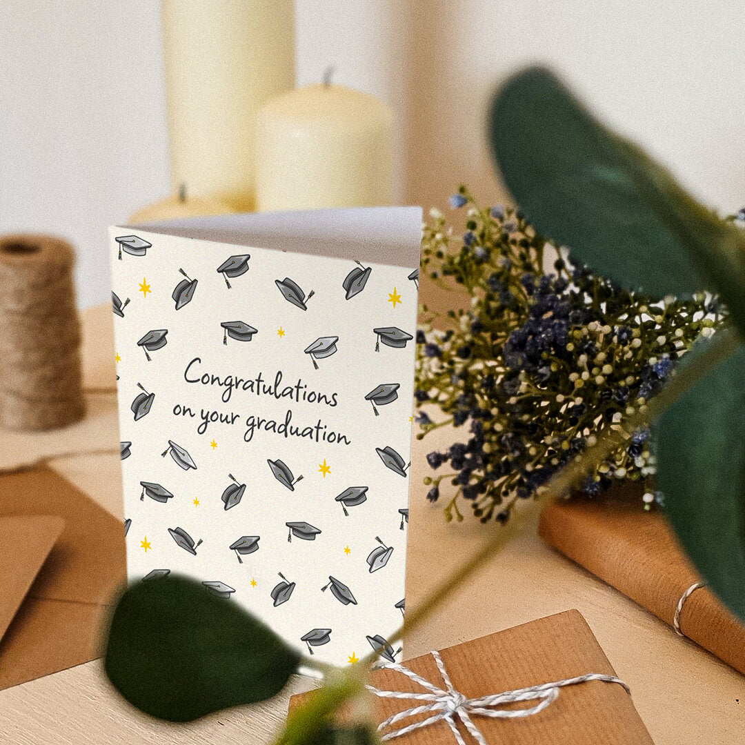 unique graduation card featuring illustrated mortarboards sat on a cream dresser with candles, gifts and plants