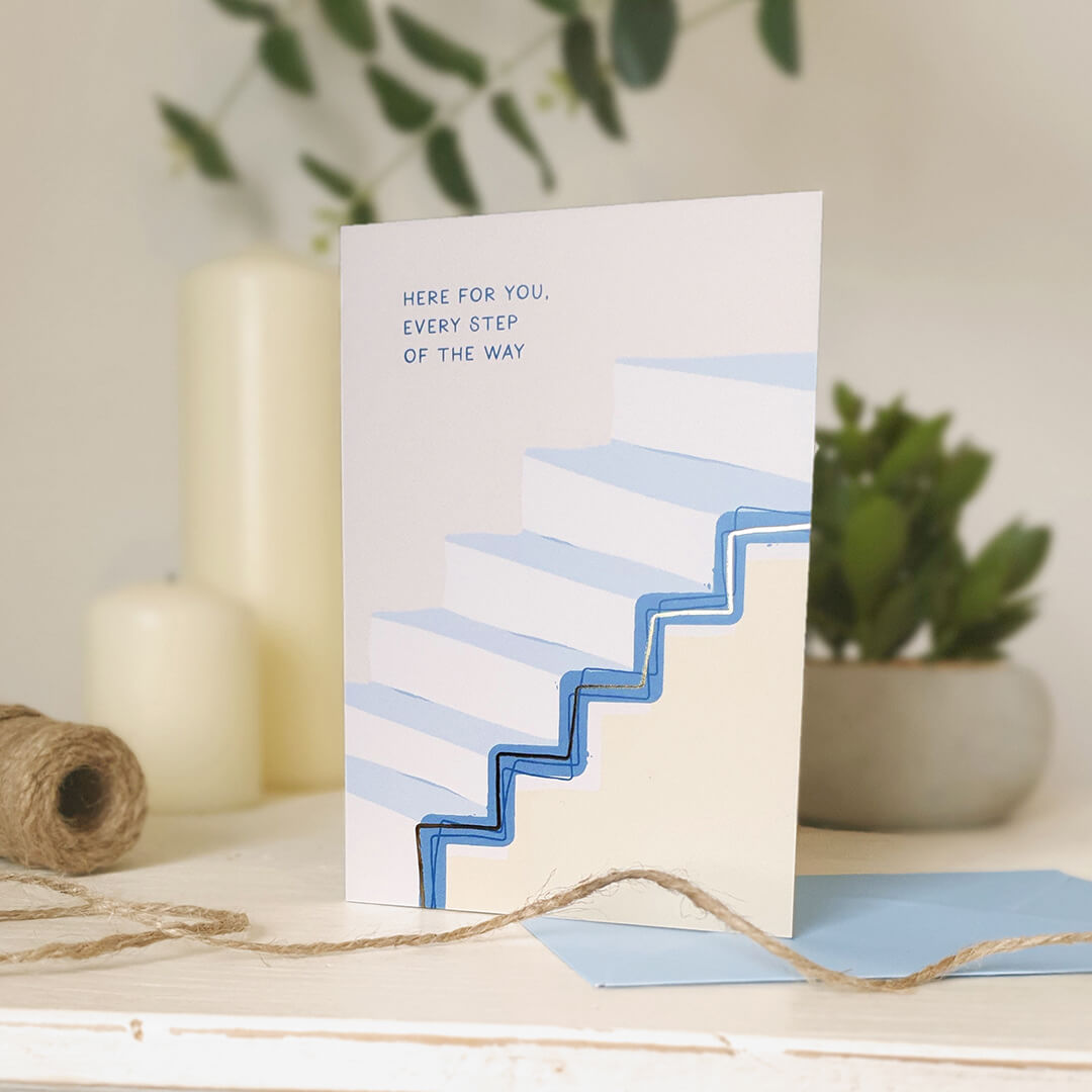 Encouragement card with blue text and staircase design, sat on a light wooden surface with plants and twine.