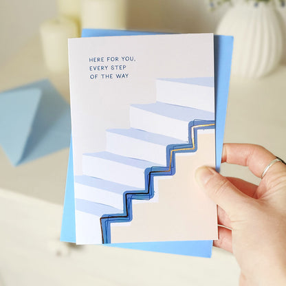 Hand holding a card with text 'Here for you, every step of the way' and a blue staircase design.