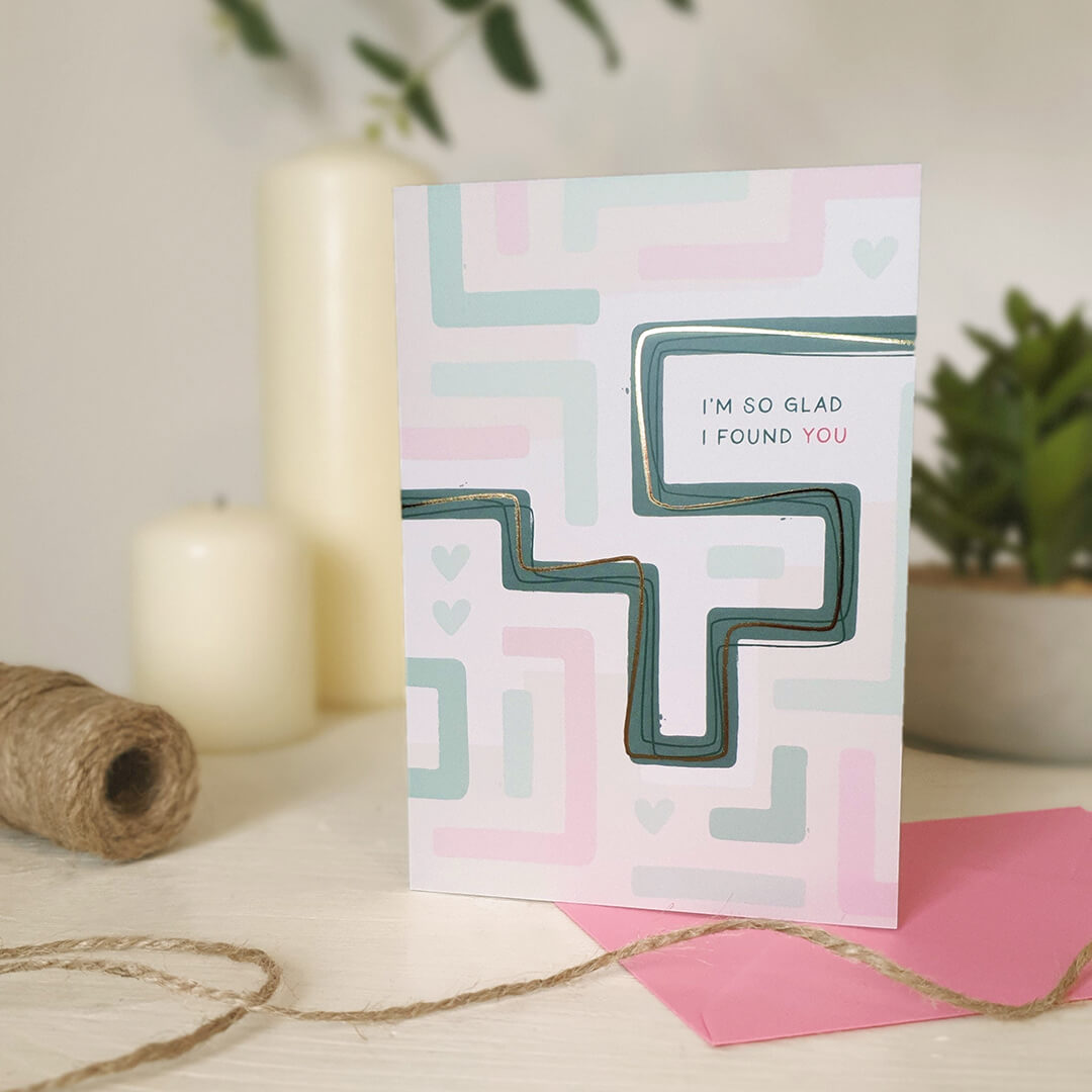 Anniversary card with geometric maze design and text sat on a light wooden dresser with candles and plants.