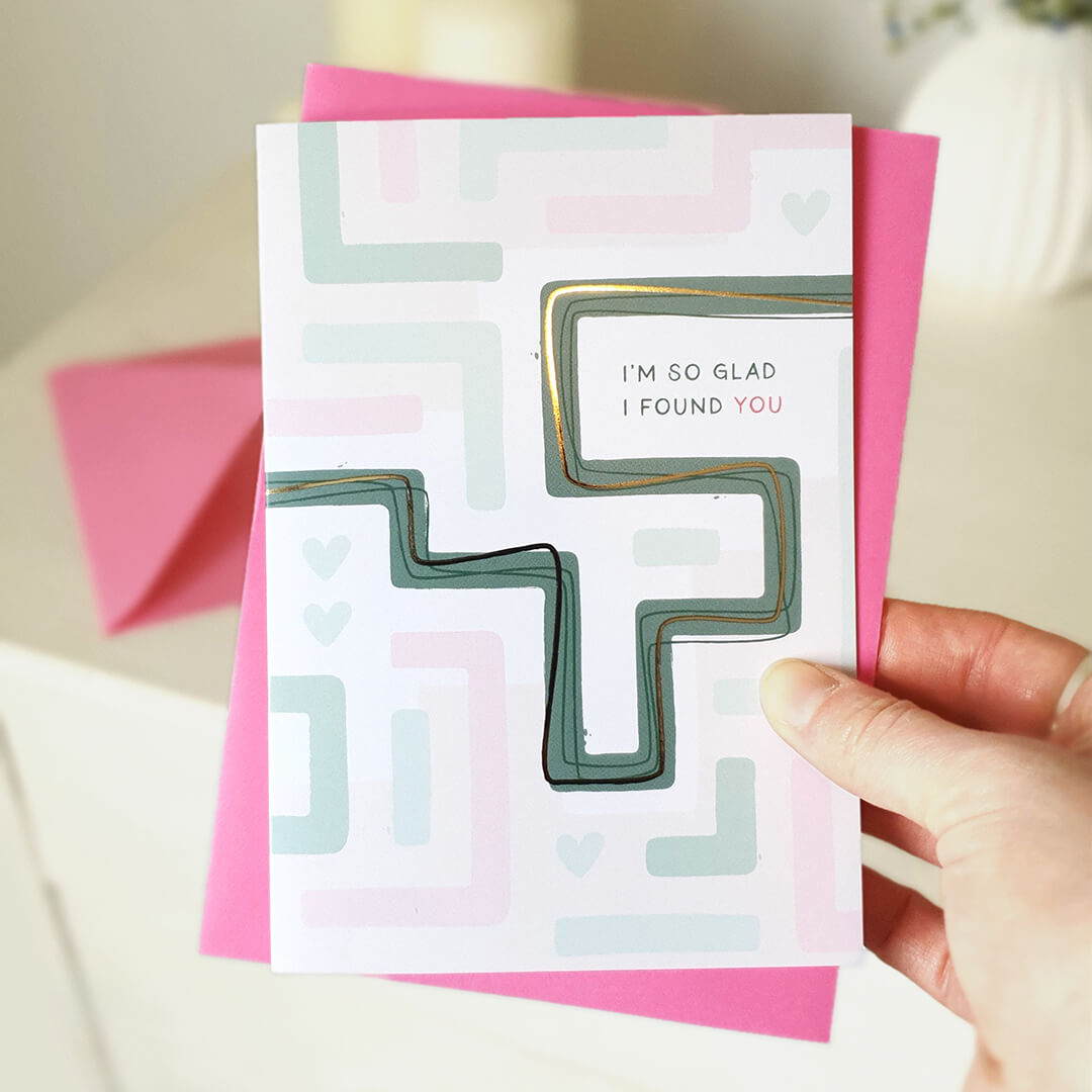 Hand holding an anniversary card with geometric maze design and text 'I'm so glad I found you' with a pink envelope.