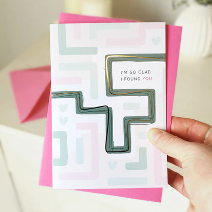 Hand holding an anniversary card with geometric maze design and text 'I'm so glad I found you' with a pink envelope.