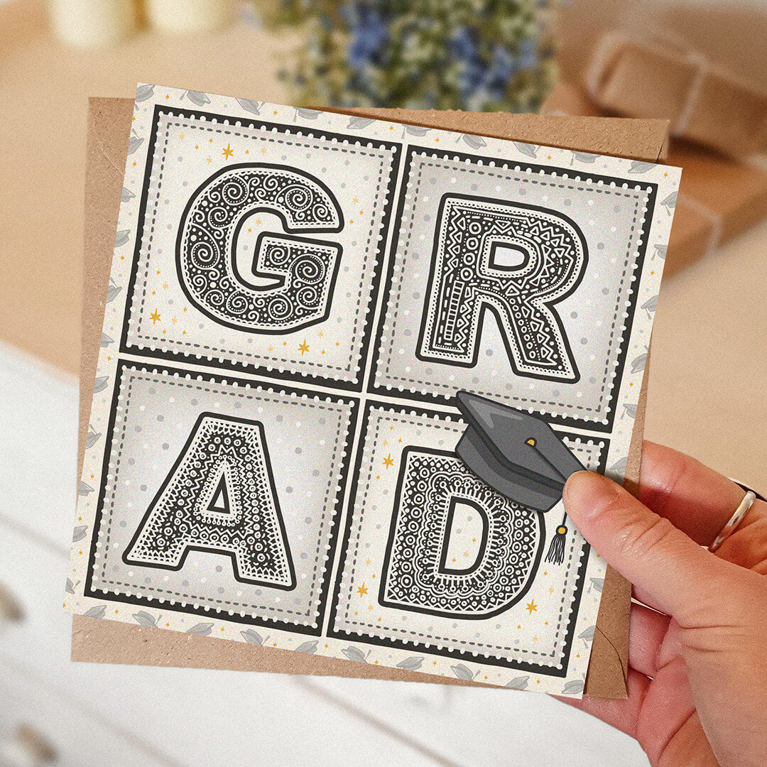 hand holding unique 'GRAD' typographic graduation card with hat illustration against a blurred background
