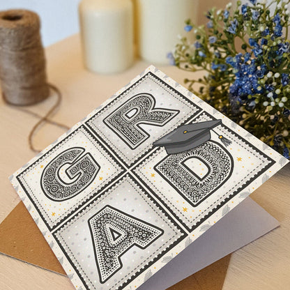 unique typographic graduation card with 'GRAD' message and hat illustration sat on a cream wooden dresser with flowers and twine in the background