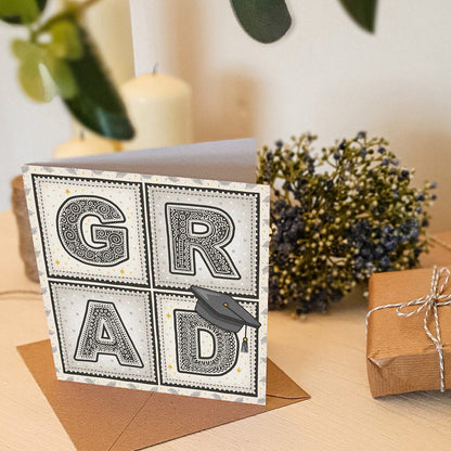 'GRAD' typographic graduation card sat on a cream wooden dresser with plants and gifts in the background