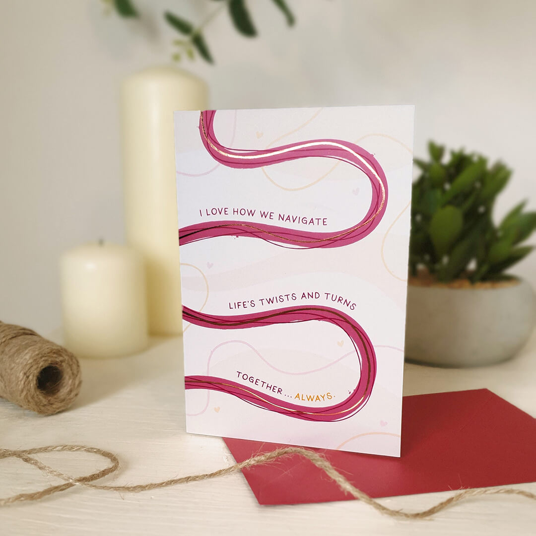 Anniversary card with pink wavy lines and text on a wooden dresser with candles and plants in the background