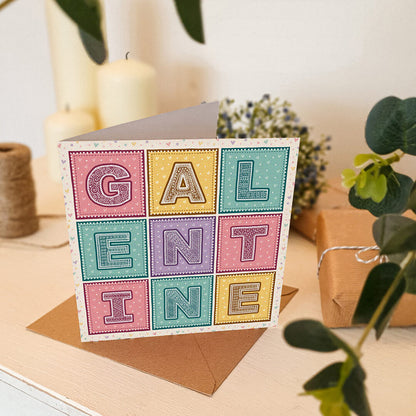 Cute pastel Galentine's Day card sat on white dresser surrounded by gifts, flowers and candles