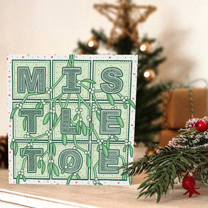 Romantic Mistletoe Christmas card with 'Mistletoe' typography and mistletoe illustrations, placed on a white wooden dresser with a blurred Christmas tree in the background.