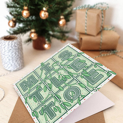 Romantic Mistletoe Christmas card with mistletoe typography and mistletoe illustrations on a white wooden dresser with wrapped gifts and a Christmas tree in the background