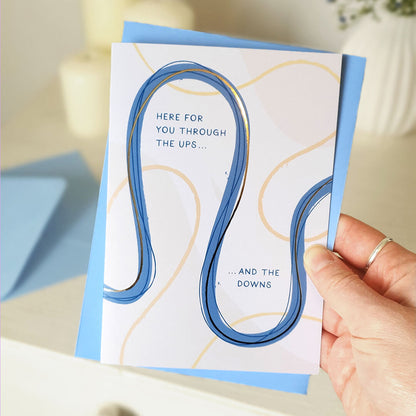 Card with blue and gold design held by a hand, with text 'Here for you through the ups... and the downs'.