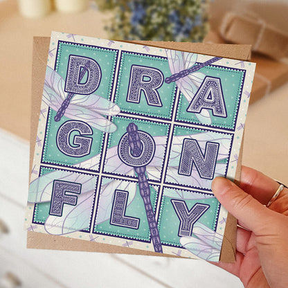 Purple and turquoise dragonfly greeting card with 'Dragonfly' text and dragonfly illustrations held by a hand on a blurred background