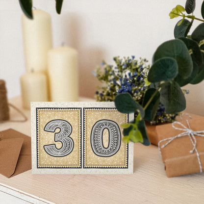 unique black and gold 30th birthday card sat on wooden dresser with candles, plants and gifts in the background