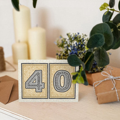 unique black and gold 40th birthday card sat on white wooden dresser with candles, plants and gifts in the background