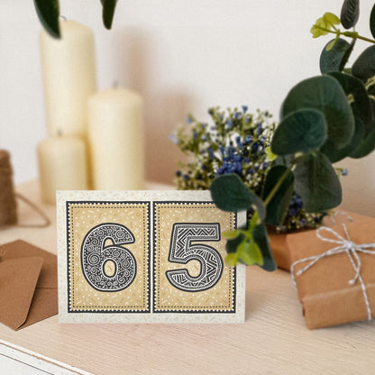 Gold 65th birthday card sat on a wooden surface with candles, plants and gifts.