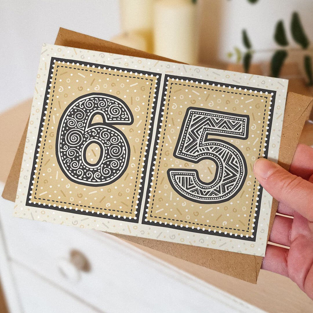 65th birthday card with decorative numbers held by a hand against a blurred neutral background