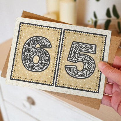 65th birthday card with decorative numbers held by a hand against a blurred neutral background