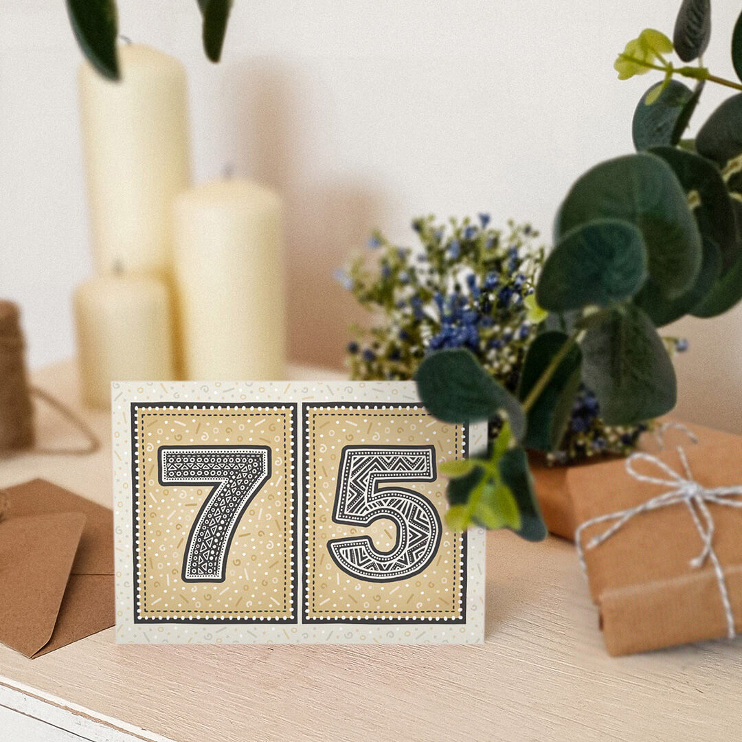 Gold 75th birthday card sat on a cream wooden dresser with candles, plants and gifts.