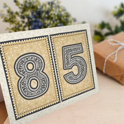 Gold 85th birthday card stood on a wooden surface with a blurred background