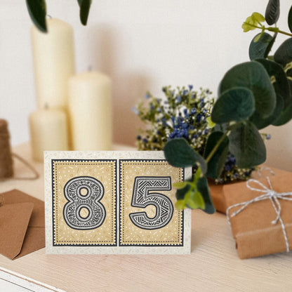 Gold 85th birthday card stood on a cream wooden table with candles, plants and gifts.