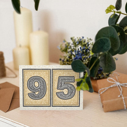 Gold 95th birthday card stood on a cream wooden surface with candles, plants and gifts.
