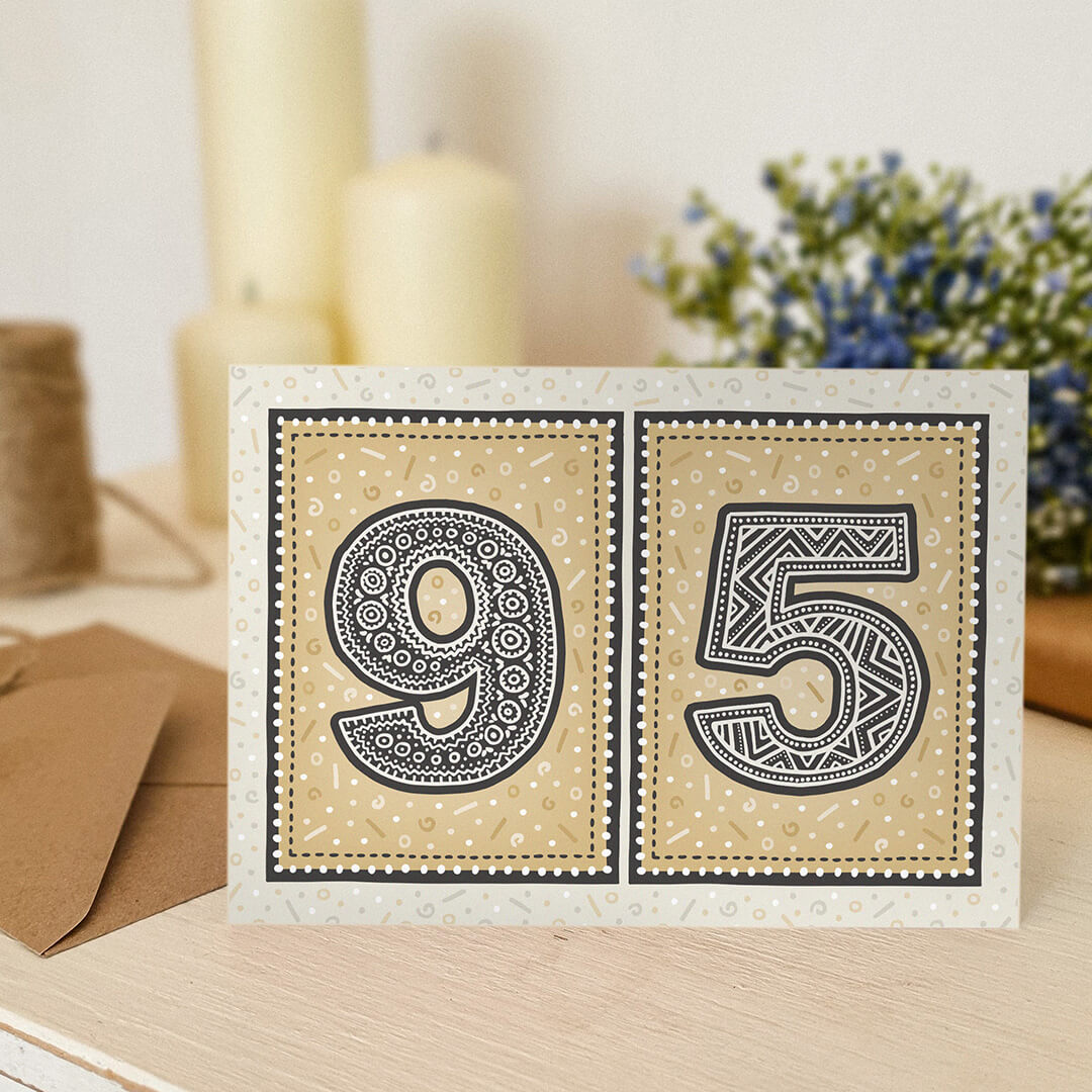 Gold 95th birthday card sat on a wooden surface with candles and flowers in the background