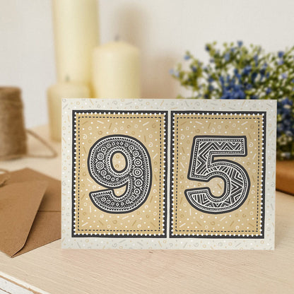 Gold 95th birthday card sat on a wooden surface with candles and flowers in the background