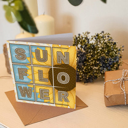 Blue and yellow sunflower greeting card with sunflower illustration on a white wooden dresser with a gift and blue flowers in the background