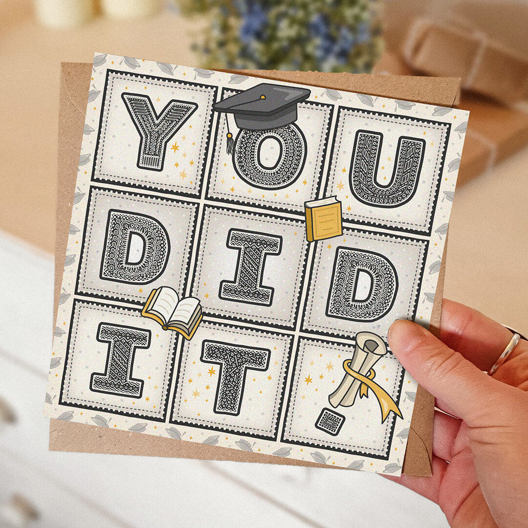 hand holding unique graduation card with 'you did it!' message and cute graduation themed illustrations with blurred background