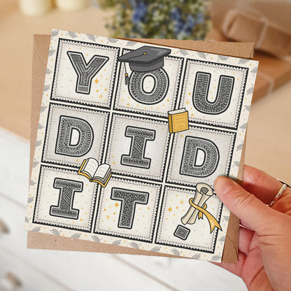 hand holding unique graduation card with 'you did it!' message and cute graduation themed illustrations with blurred background