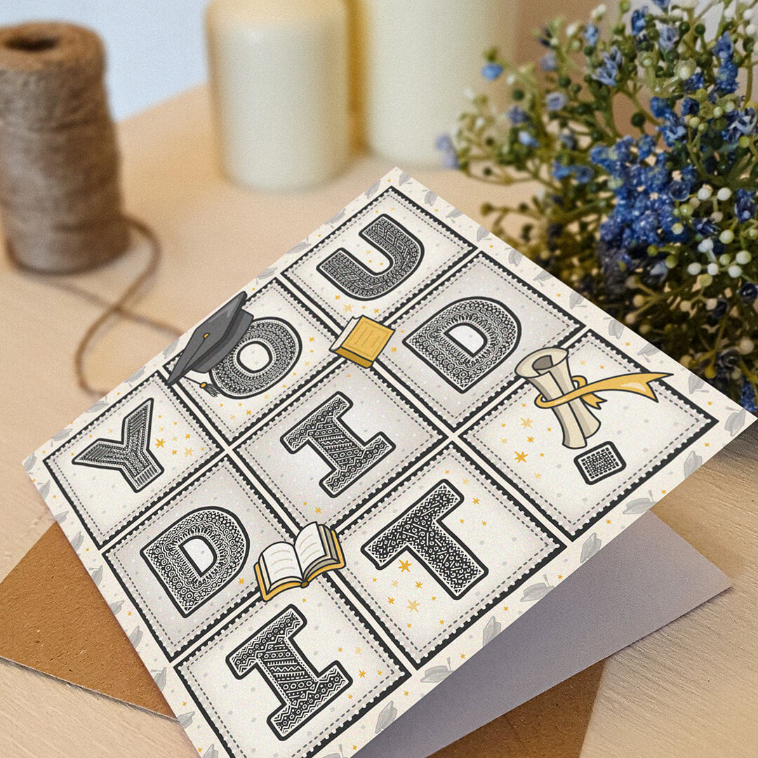 unique 'you did it!' graduation card with scroll, hat and book illustrations sat on a cream wooden dresser with flowers and twine in the background