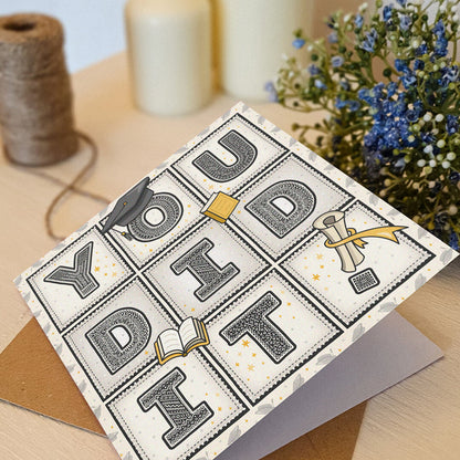 unique 'you did it!' graduation card with scroll, hat and book illustrations sat on a cream wooden dresser with flowers and twine in the background