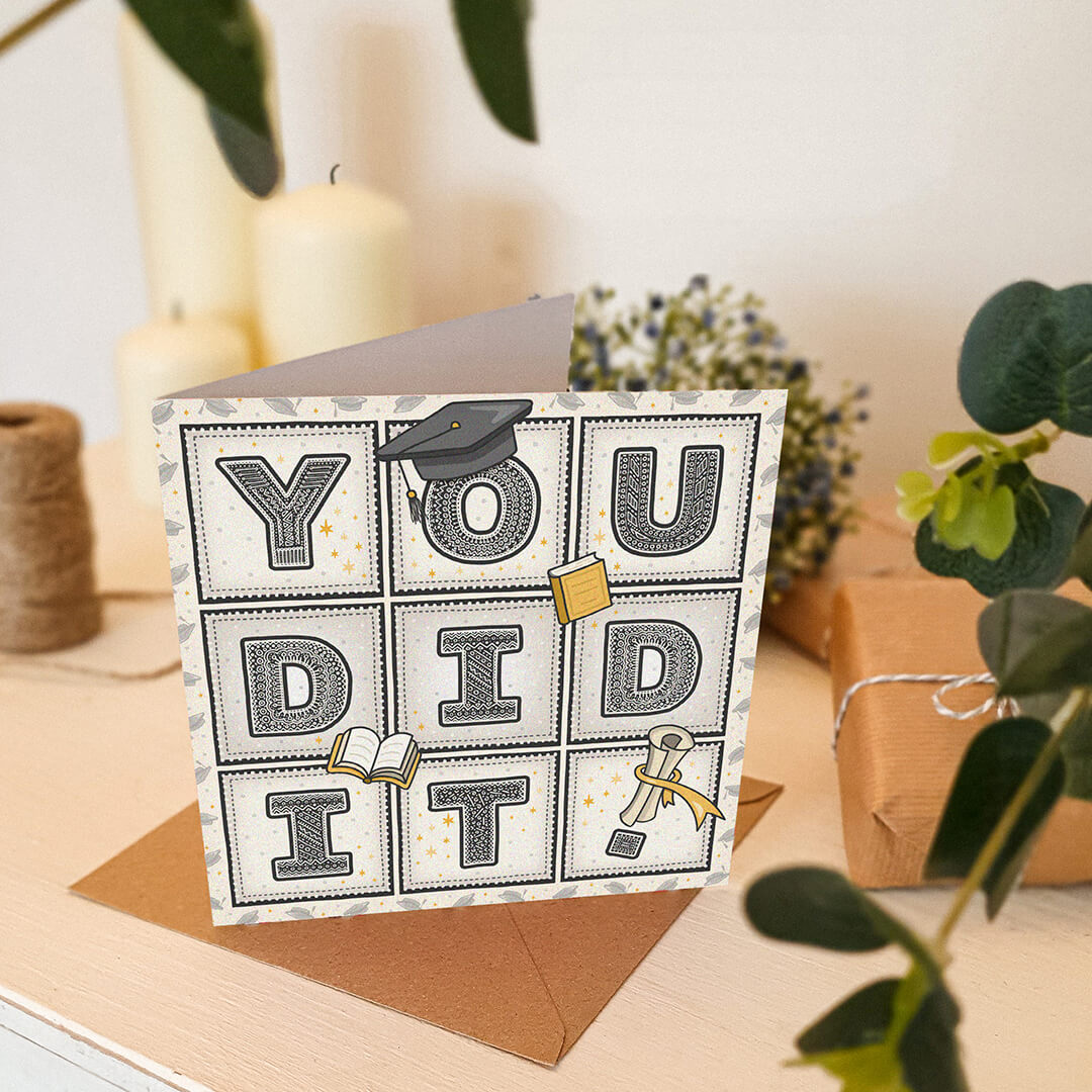 unique graduation card with 'you did it!' message and scroll, hat and book illustrations sat on a cream wooden dresser with gifts, plants and candles in the background