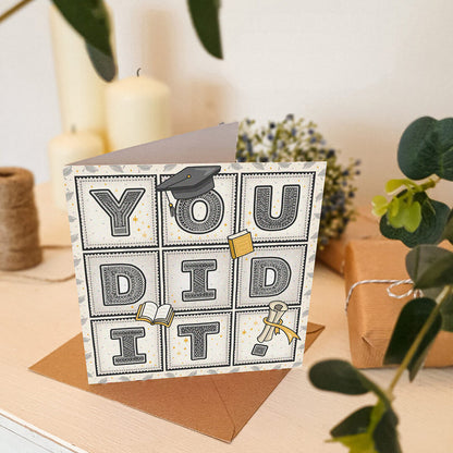 unique graduation card with 'you did it!' message and scroll, hat and book illustrations sat on a cream wooden dresser with gifts, plants and candles in the background